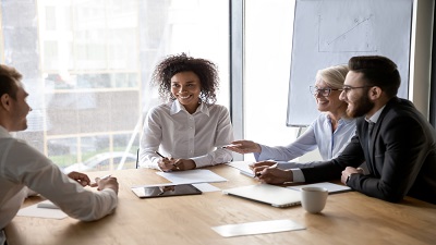 Four people seated around a wooden conference table in a modern office, engaged in a discussion. The table holds papers, notebooks, a smartphone, and a coffee cup. A large window lets in natural light, and a flip chart with graphs is visible in the background.