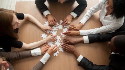 A group of people seated around a wooden table, each reaching toward the center where several white puzzle pieces are laid out, symbolizing collaboration and teamwork.