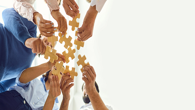 A group of people collaboratively holding and connecting large wooden puzzle pieces, with their hands reaching toward the center to fit the pieces together.